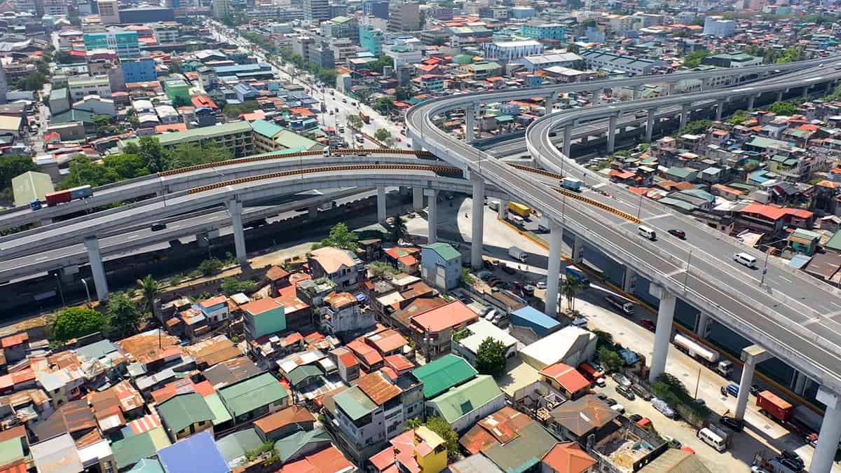 Hình chụp trên không một nút giao thông nhiều tầng phía trên khu đô thị đông dân, với mái nhà đa màu sắc và đủ loại phương tiện di chuyển trên hạ tầng được số hóa bên dưới — dự án NLEX-SLEX Connector Road giúp giảm tắc nghẽn cho khu vực chiếm 36% kinh tế Manila.