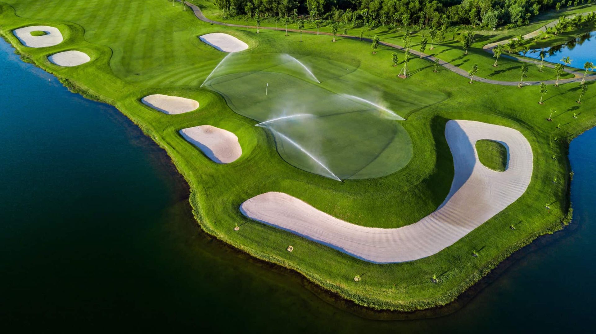 Toàn cảnh sân golf với hệ thống tưới nước trên cỏ, bao quanh là bẫy cát, hồ nước và cây xanh, lồng ghép hoàn hảo với khu dân cư cạnh đó.