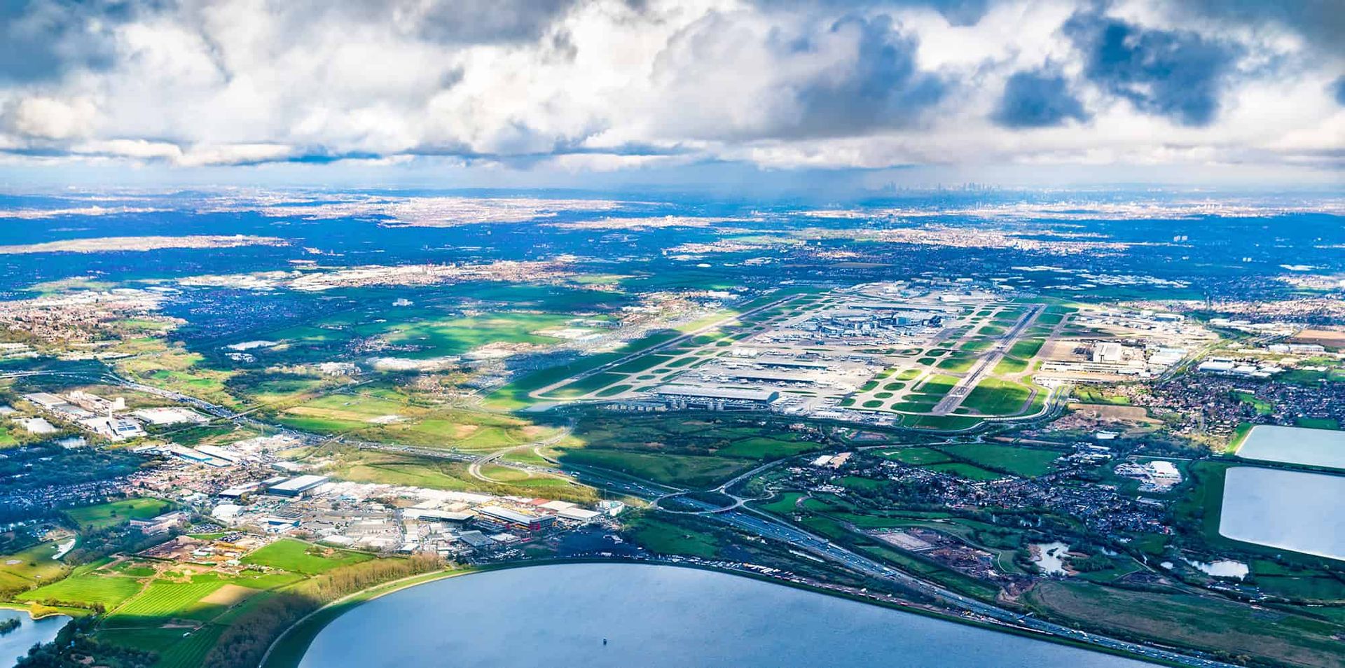 Toàn cảnh trên cao sân bay Heathrow ở London, Anh, với các đường băng, nhà ga, cùng những cánh đồng xanh và kiến trúc lân cận trời mây nhẹ.